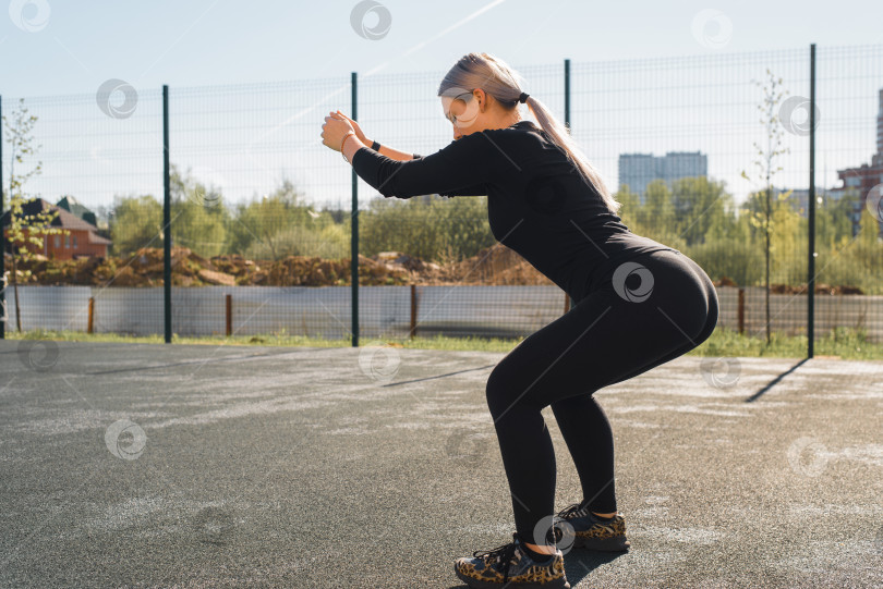 Скачать Уличная тренировка для ягодиц и ног. Молодая женщина, занимающаяся фитнесом, делает приседания, утро на свежем воздухе. Спортсменка в черном облегающем спортивном костюме потягивается на детской площадке. Здоровый активный образ жизни фотосток Ozero