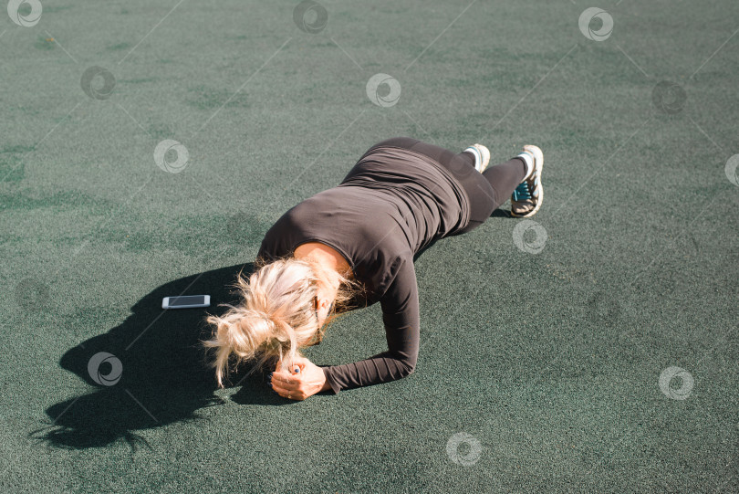 Скачать Женщина занимается тренировкой на открытом воздухе. Спортсменка в обтягивающем спортивном костюме некоторое время выполняет упражнение на доске по мобильному телефону. Тренировка на спортивной площадке летним днем. Потеря веса, концепция активного образа жизни фотосток Ozero