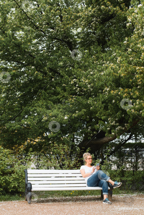 Скачать Женщина отдыхает в парке на скамейке и пользуется мобильным телефоном. Улыбающаяся дама общается с кем-то в мессенджере на смартфоне на открытом воздухе. Вертикальная фотография фотосток Ozero