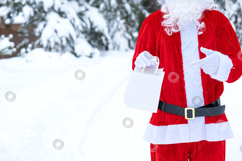 Скачать Санта-Клаус на открытом воздухе зимой и в снегу, раздающий в руки бумажные пакеты, указывает пальцем. Покупки, переработка упаковки, ручная работа, доставка на Рождество и Новый год фотосток Ozero
