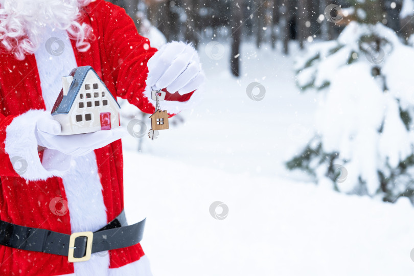 Скачать Ключ от дома с брелком коттедж в руках Деда Мороза на открытом воздухе в снегу. Сделка с недвижимостью, покупка, строительство, переезд, ипотека. Уютный дом. Счастливого Рождества, нового года, бронирования мероприятий и зала фотосток Ozero