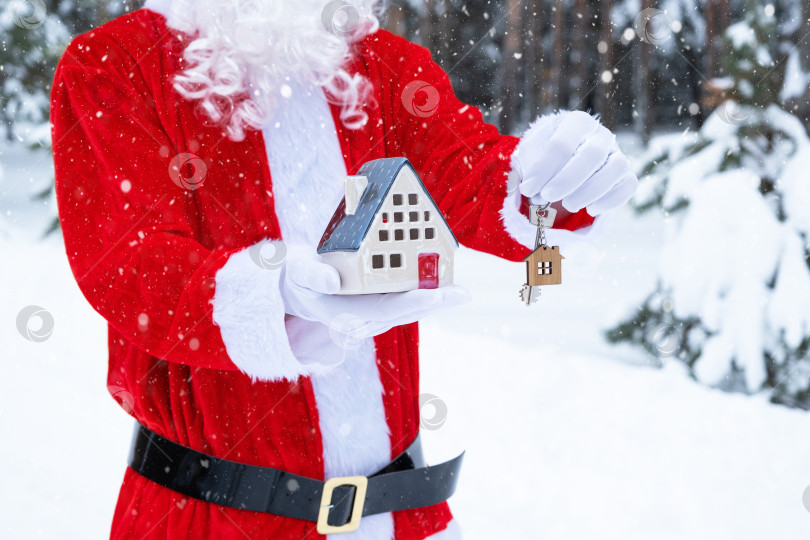 Скачать Ключ от дома с брелком коттедж в руках Деда Мороза на открытом воздухе в снегу. Сделка с недвижимостью, покупка, строительство, переезд, ипотека. Уютный дом. Счастливого Рождества, нового года, бронирования мероприятий и зала фотосток Ozero