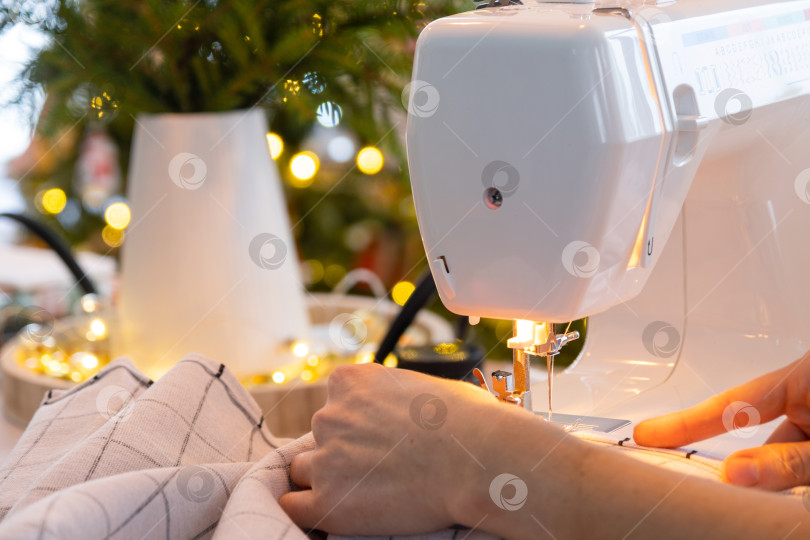 Скачать Процесс шитья на швейной машинке, женские руки управляют иглой с ниткой из белой клетчатой ткани. Подготовка к Рождеству и Новому году, текстиль для уюта в праздничном декоре интерьера фотосток Ozero