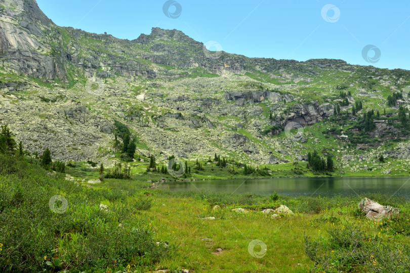 Скачать Пологий берег красивого озера, заросшего травой и кустарником, окруженный высокими горными хребтами в солнечный летний день. фотосток Ozero