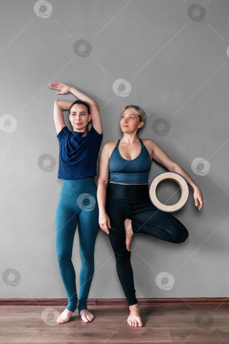 Скачать Обрадованные женщины-тренеры по йоге в полный рост в спортивной одежде стоят босиком у стены, одна держит в руке деревянный круг, радуясь началу занятий или расслабляясь после окончания урока фотосток Ozero
