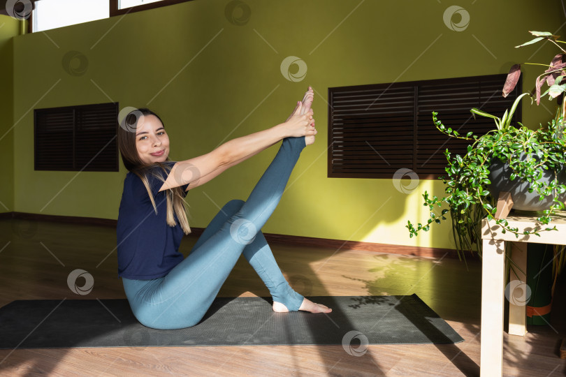 Скачать Молодая улыбчивая спортсменка-шатенка выполняет динамическую растяжку мышц, сгибающих ее ногу в колене, тренируется в спортивной одежде, сидя на коврике в современной фитнес-студии фотосток Ozero