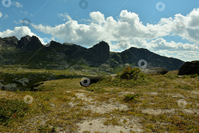 Скачать Вид с вершины скалистого холма, поросшего кустарником, на высокий горный хребет с вершинами под облачным летним небом. фотосток Ozero