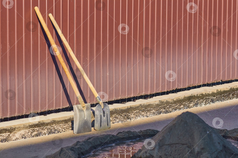 Скачать Две лопаты стоят у стены, сделанной из металлического профилированного железа. Смесь цемента и песка замачивают в воде, чтобы замесить бетон вручную. фотосток Ozero