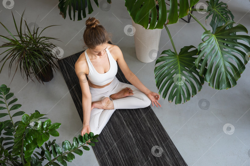 Скачать Женщина, ведущая здоровый образ жизни и практикующая йогу, занимающаяся медитацией, сидящая в позе лотоса с джняна мудрой на коврике, окруженном различными цветами, тренирующаяся в белой спортивной одежде в комнате фотосток Ozero