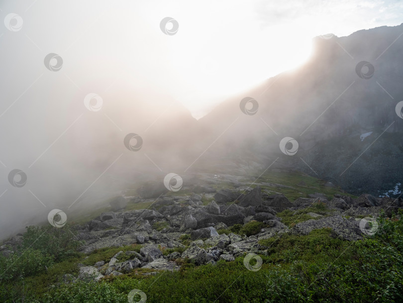 Скачать Красочный зеленый пейзаж с камнями на холме на фоне гигантской горной стены в туманном солнечном свете. Минималистичный яркий солнечный пейзаж с солнцем и солнечными вспышками. Минималистичный альпийский вид. Живописный минимализм фотосток Ozero