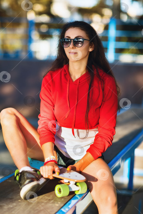 Скачать Молодая женщина в солнцезащитных очках отдыхает на спортивной площадке для катания на скейтборде летним солнечным вечером фотосток Ozero