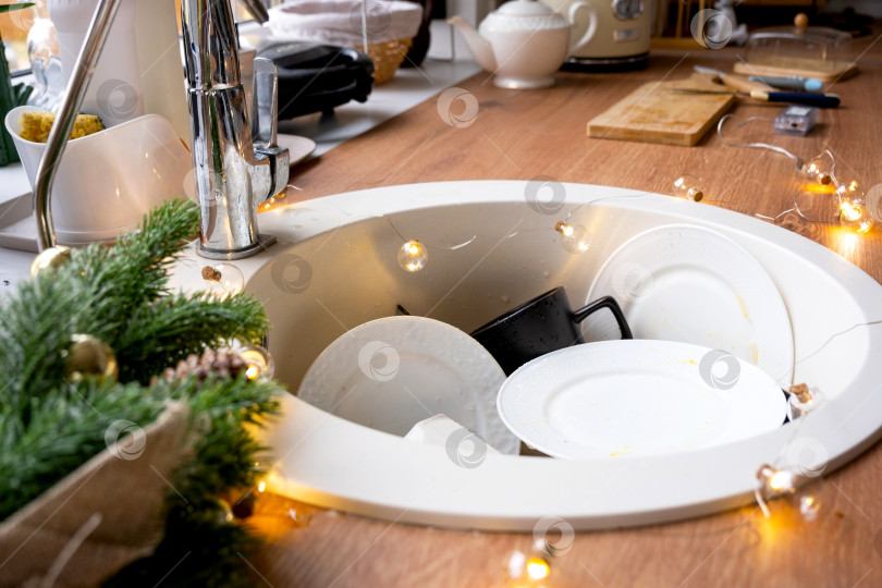 Скачать Грязная посуда в раковине на украшенной гирляндами кухне, праздник после рождественского застолья. Уборка после гостей на Новый год, уборка кухни, моющие средства, клининговые услуги фотосток Ozero