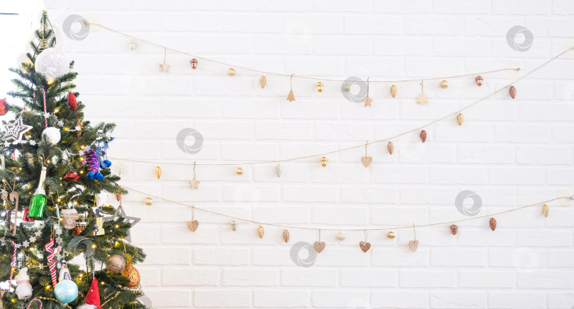 Скачать Рождественская елка у белой кирпичной стены с гирляндой из стеклянных игрушек в форме буквы Z на веревке. Светящиеся гирлянды украшают комнату-студию. Уютный дом на Рождество и новый год фотосток Ozero