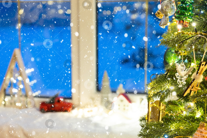 Скачать Уютный рождественский декор с домиками, грузовичком на подоконнике и снегопадом возле рождественской елки. фотосток Ozero