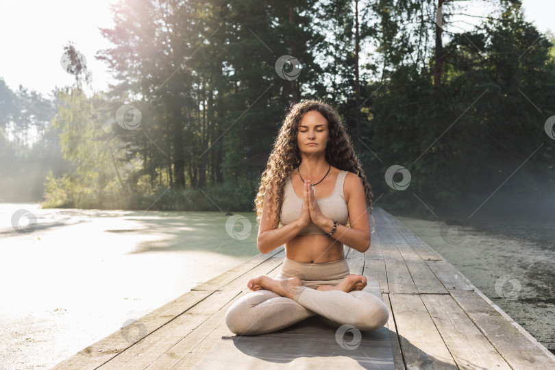 Скачать Женщина, практикующая йогу, выполняющая упражнение падмасана с намасте, медитирующая, сидя в позе лотоса на деревянном мосту в парке летним утром фотосток Ozero