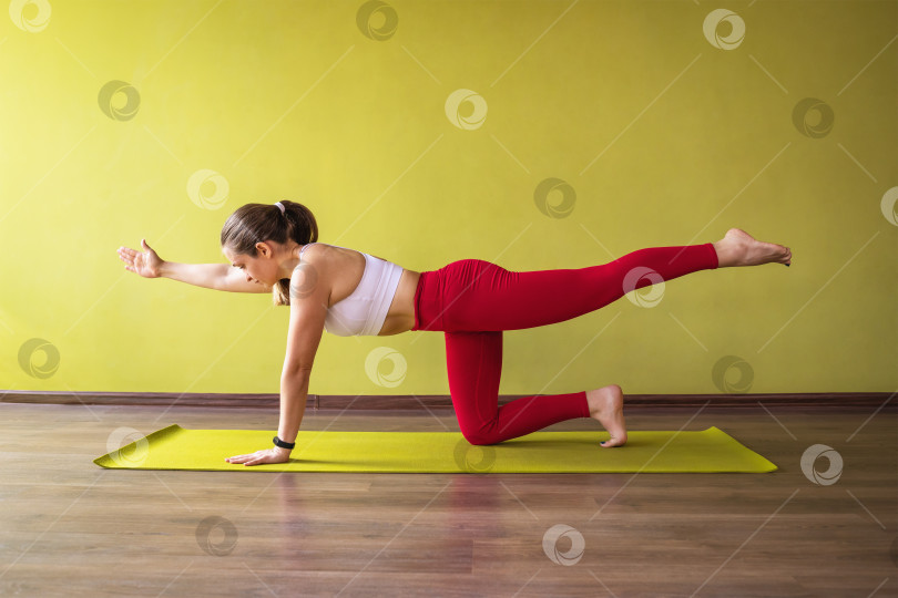 Скачать Женщина, практикующая йогу, выполняющая вариацию упражнения Паршва Марджариасана, поза журавля, тренируется в студии у стены на коврике фотосток Ozero