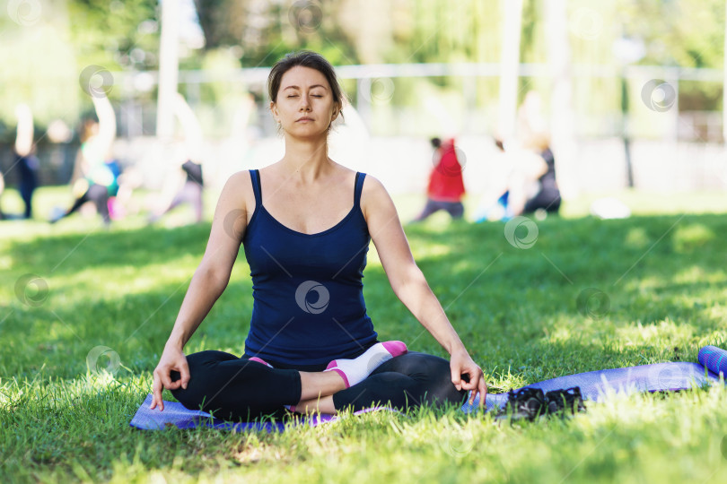 Скачать Женщина, ведущая здоровый образ жизни и практикующая йогу, медитирует, сидя в позе лотоса на траве солнечным летним утром в парке на фоне группы людей, занимающихся спортом фотосток Ozero