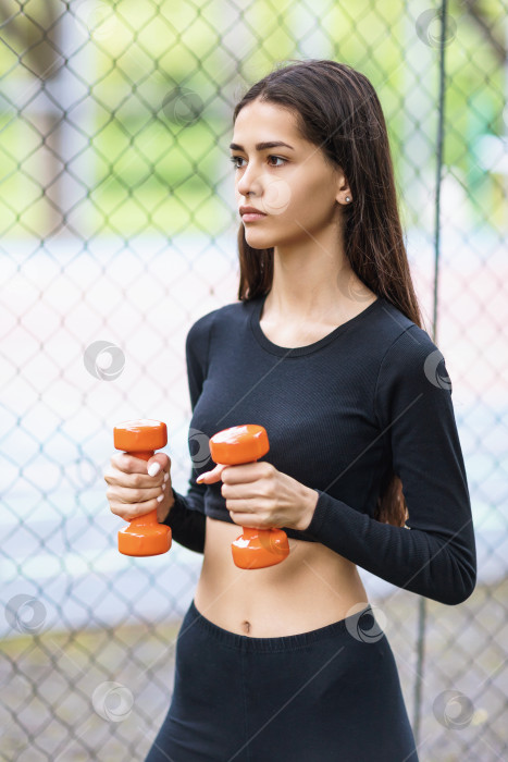 Скачать Молодая красивая девушка, ведущая здоровый образ жизни, тренируется в спортивной одежде на улице с гантелями фотосток Ozero