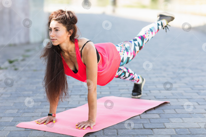 Скачать Молодая женщина, ведущая здоровый образ жизни, тренируется летом на набережной в спортивной одежде, выполняет фитнес-упражнения на коврике фотосток Ozero