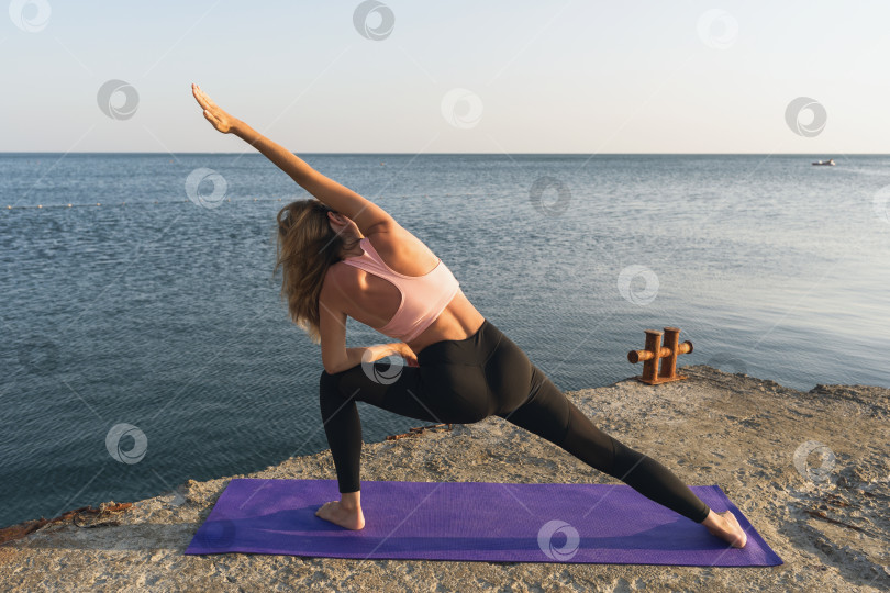 Скачать Женщина в спортивной одежде, практикующая йогу, выполняет упражнение уттхита паршваконасана на берегу моря, стоя на коврике, угловая поза с растяжкой фотосток Ozero