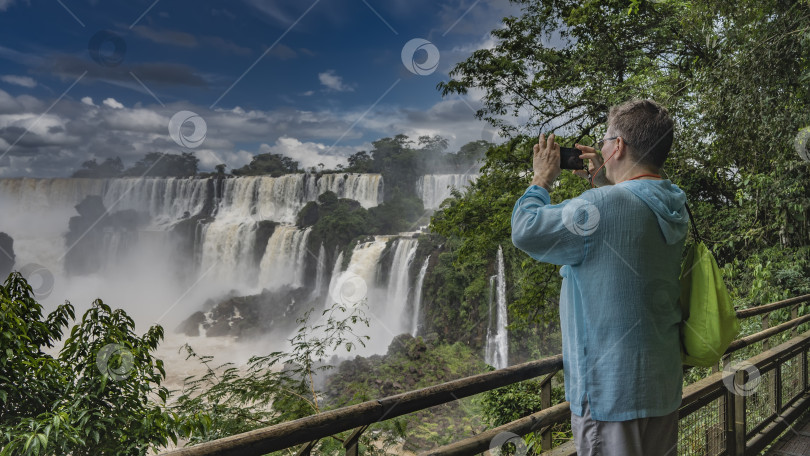 Скачать Мужчина стоит на смотровой площадке у перил, фотографируя водопад на свой смартфон. Рюкзак на плече. Потоки воды срываются с выступов скал. Брызги, туман. фотосток Ozero