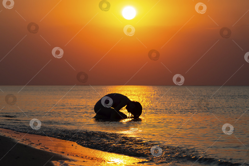 Скачать Силуэт девочки-подростка в морской воде возле песчаного пляжа во время ярко-оранжевого заката, мягкий фокус. Девушка наслаждается купанием на фоне удивительного заката. концепция ухода за телом фотосток Ozero