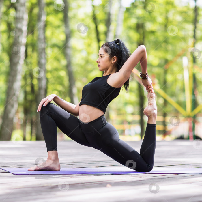 Скачать Молодая девушка, практикующая йогу, выполняет вариацию упражнения эка пада раджакапотасана, позу короля голубей, тренируется в парке на деревянном мосту в черной спортивной одежде фотосток Ozero