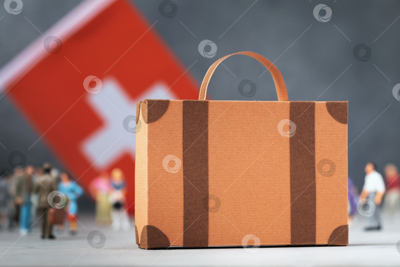 Скачать Картонный чемодан, пластмассовые игрушечные человечки и флаг на абстрактном фоне, концепция на тему переезда или миграции в Швейцарии фотосток Ozero