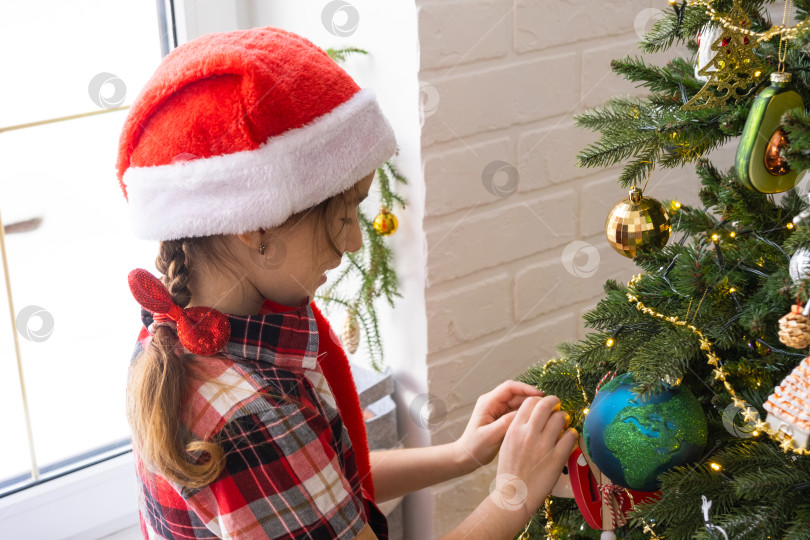 Скачать Счастливая девочка в шляпе Санты украшает рождественскую елку разноцветными фигурками. Ребенок веселится босиком, в колпаке гнома у окна, в ожидании Рождества и Нового года фотосток Ozero