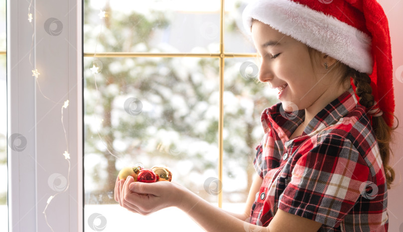Скачать Девушка в шапке Санты сидит на подоконнике дома возле рождественской елки и держит в руках стеклянные шары. Ребенок веселится, верит в чудо и ждет Рождества и Нового года фотосток Ozero