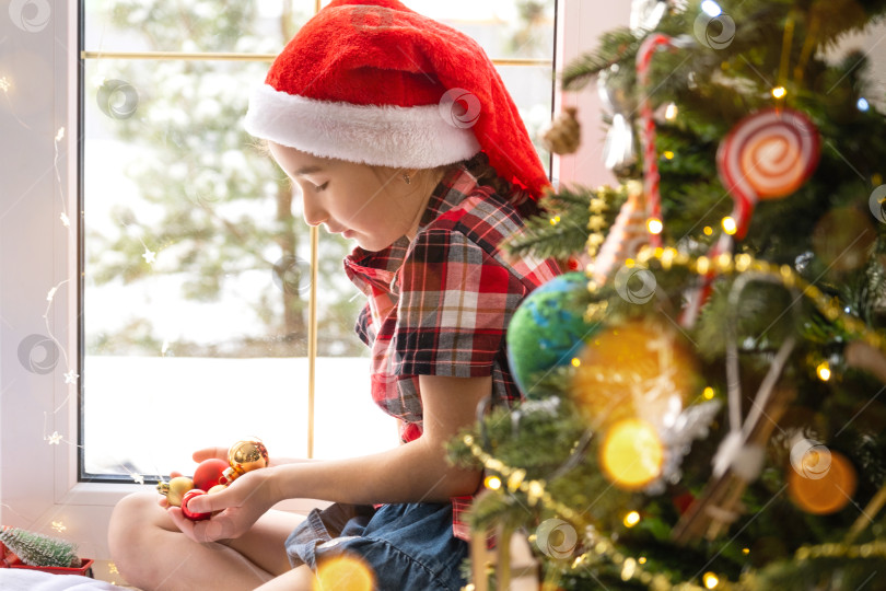 Скачать Девушка в шапке Санты сидит на подоконнике дома возле рождественской елки и держит в руках стеклянные шары. Ребенок веселится, верит в чудо и ждет Рождества и Нового года фотосток Ozero