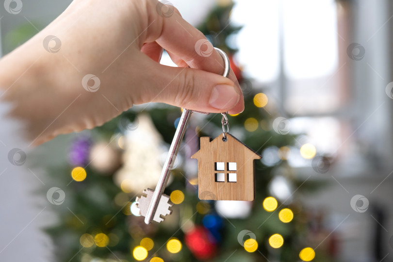 Скачать Ключ и брелок с крошечным домиком в руке на фоне рождественской елки с гирляндами. подарок на Новый год, Рождество. Строительство, дизайн, проект, переезд, страхование, ипотека, аренда и покупка недвижимости. фотосток Ozero