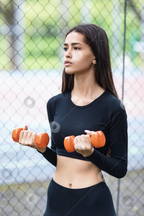 Скачать Молодая девушка, ведущая здоровый образ жизни, тренируется в спортивной одежде с гантелями на улице фотосток Ozero