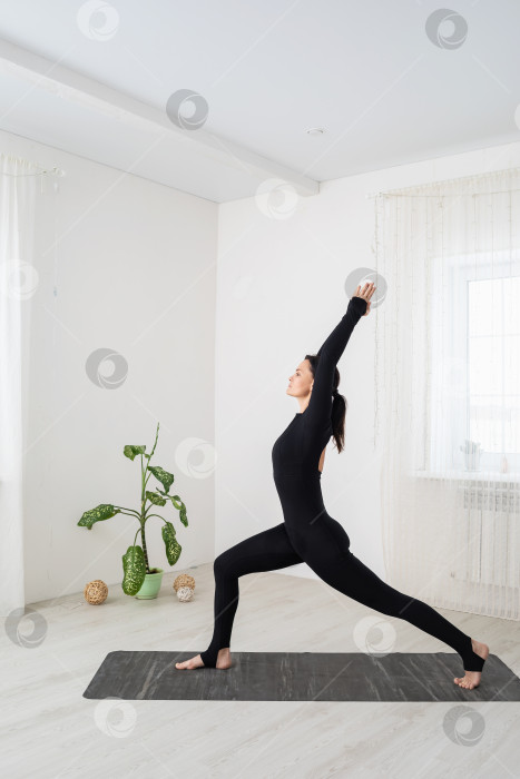 Скачать Женщина, ведущая здоровый образ жизни и практикующая йогу, выполняет упражнение вирабхадрасана, позу воина, тренируется в черной спортивной одежде, стоя на коврике в комнате фотосток Ozero