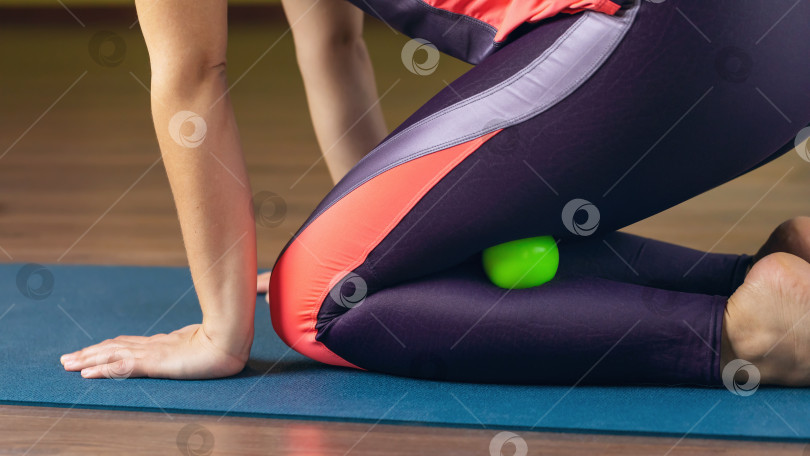 Скачать Женщина выполняет миофасциальную релаксацию сверхгибких мышц ног с помощью массажного мяча, сидя на коврике в фитнес-студии и тренируясь в спортивной одежде фотосток Ozero