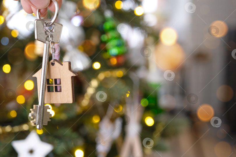 Скачать Ключ и брелок с крошечным домиком в руке на фоне рождественской елки с гирляндами. подарок на Новый год, Рождество. Строительство, дизайн, проект, переезд, страхование, ипотека, аренда и покупка недвижимости. фотосток Ozero