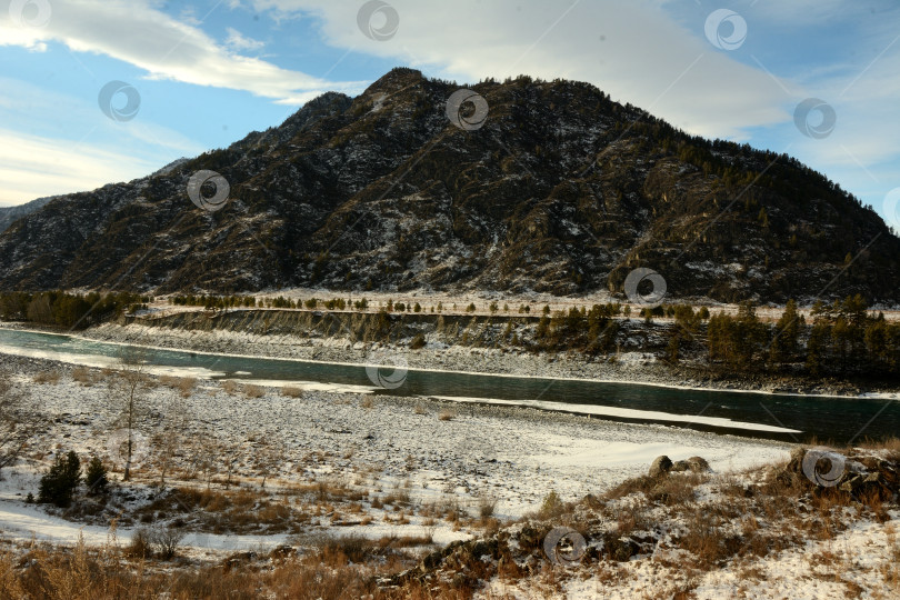 Скачать Вид со скалистого берега красивой реки с песчаными берегами, покрытыми снегом, текущей вдоль высоких гор солнечным зимним днем. фотосток Ozero