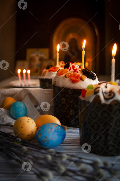 Скачать Пасхальный натюрморт с пасхальным куличом, пасхальными яйцами, ветками вербы и горящими свечами в темноте монастырской церкви с русскими иконами на заднем плане фотосток Ozero