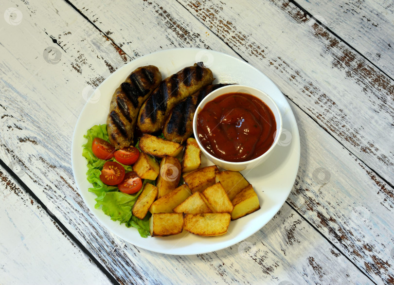 Скачать Тарелка с вкусным обедом, сосиски-гриль с картофелем, помидорами, листьями салата и соусом на светлом деревянном столе. фотосток Ozero