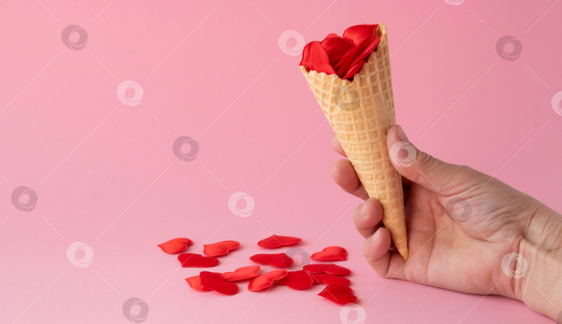 Скачать В руке рожок мороженого с красными сердечками. Креативная фотография пустых вафельных рожков с разбросанными красными сердечками на розовом фоне. Понятие любви, лета, весны. место для текста фотосток Ozero