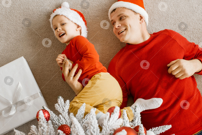 Скачать Вид сверху на счастливую радостную семью: папа и сын в шапочках Санты играют и веселятся на полу под рождественской елкой. Празднование Рождества дома. фотосток Ozero