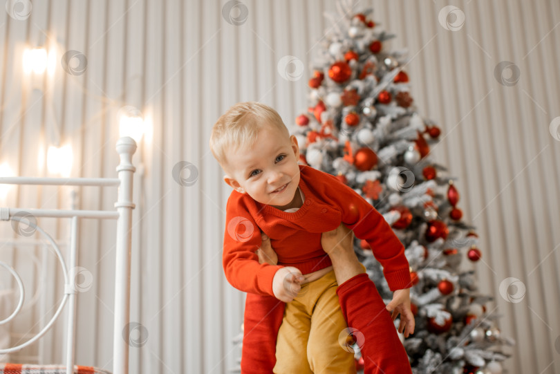 Скачать Милый улыбающийся маленький сын в красном свитере веселится на руках у отца и указывает пальцем в камеру на фоне рождественской елки фотосток Ozero