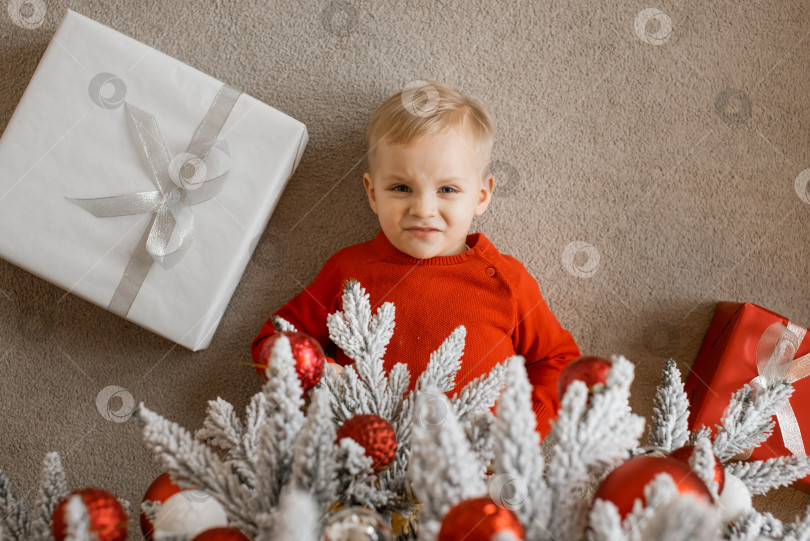 Скачать Вид сверху на смеющегося светловолосого маленького мальчика в красном вязаном свитере с кучей рождественских подарков, лежащих под украшенной елкой фотосток Ozero