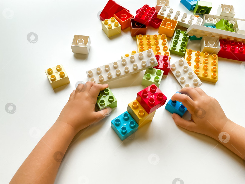Скачать Ребенок играет с разноцветными строительными блоками на белом фоне. Вид сверху фотосток Ozero