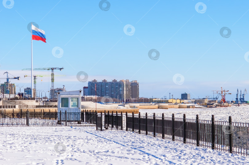 Скачать Большой российский флаг на заснеженной набережной реки Амур солнечным зимним днем. Гранитный прибрежный комплекс в городе Благовещенске, Россия. Речной порт и строительные краны фотосток Ozero