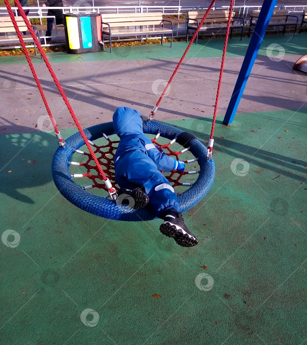 Скачать Маленький мальчик в синем комбинезоне качается на качелях на детской площадке весной или осенью. фотосток Ozero