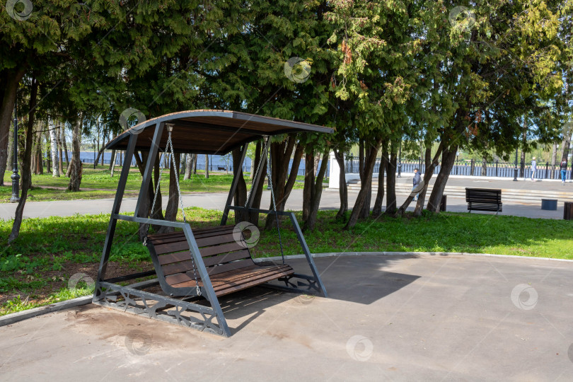 Скачать Деревянная коричневая скамейка-качели в городском парке ярким весенним днем фотосток Ozero