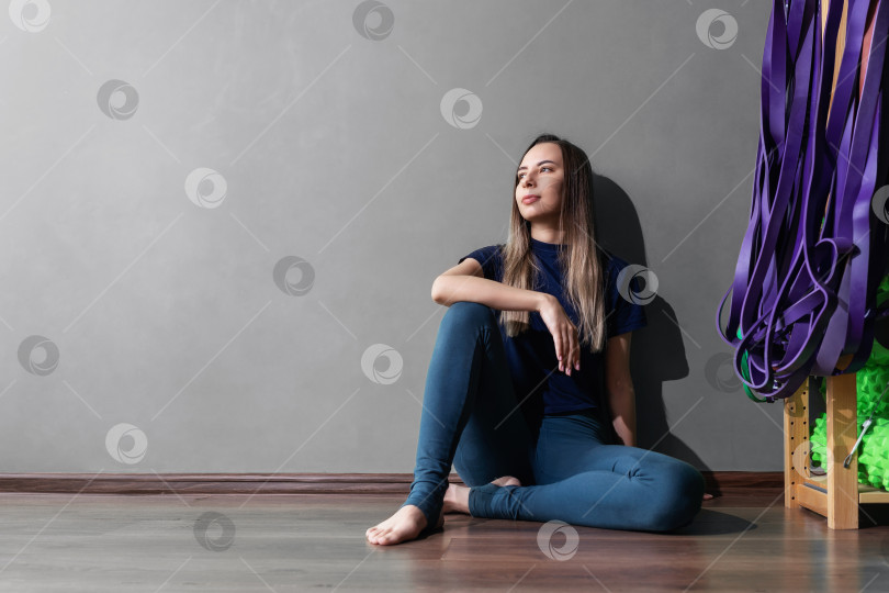 Скачать Молодая женщина отдыхает после тренировки по фитнесу, сидит спиной к стене в спортивной одежде, портрет в полный рост с копией пространства фотосток Ozero