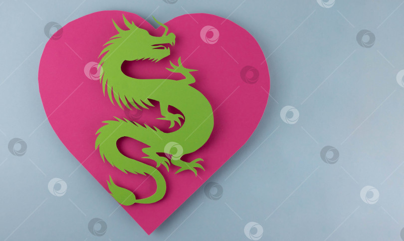 Скачать Зеленый бумажный дракон лежит на розовом сердечке на синем фоне. Концепция нового 2024 года фотосток Ozero