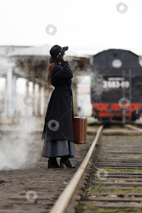 Скачать Красивая девушка в черном пальто и шляпе с чемоданом в руках возле старого паровоза. Молодая женщина с длинными темными волосами. Винтажный портрет прошлого века, ретро-путешествие. фотосток Ozero